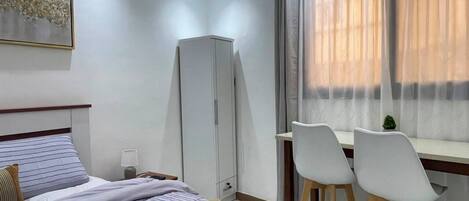 A bedroom with a bed, a desk, two chairs, a wardrobe, and a window with curtains.