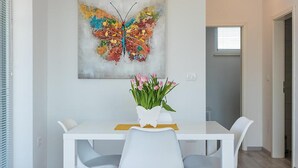 Un comedor con una mesa blanca, dos sillas, un florero con flores y un cuadro de una mariposa colorida en la pared.