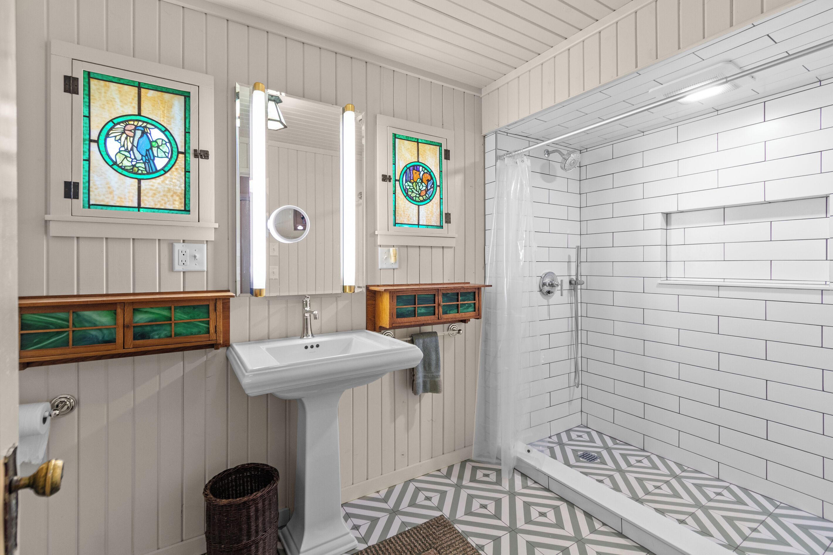 Une salle de bain avec un lavabo blanc, deux fenêtres à vitraux, une douche aux carreaux blancs et un sol à motifs.