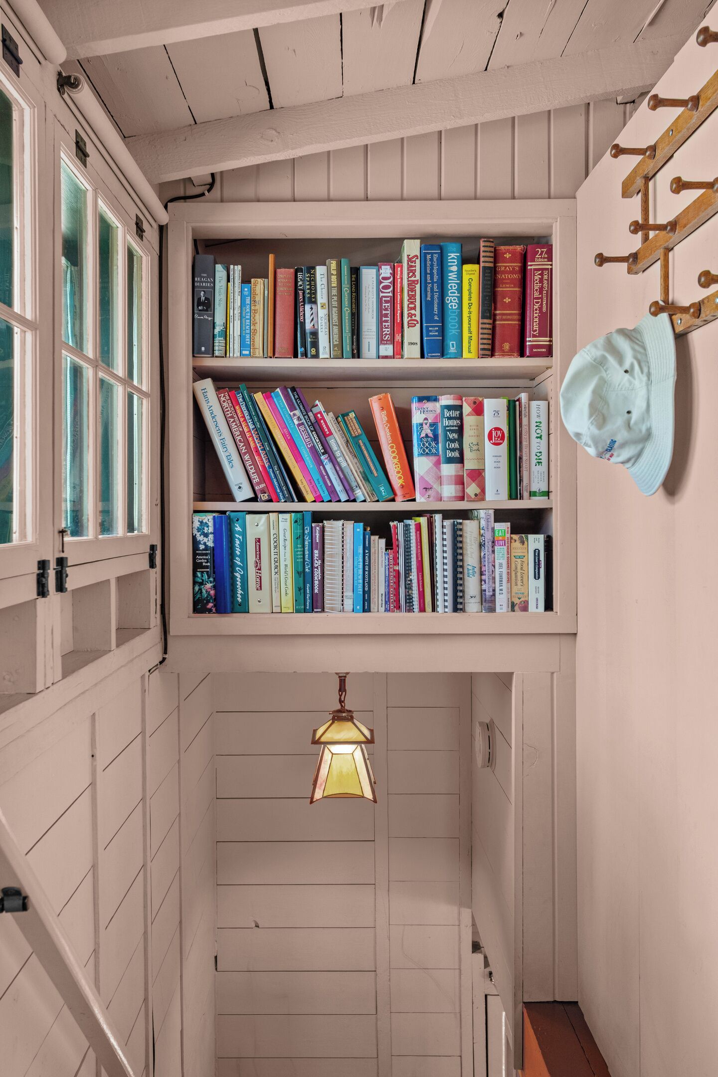 A narrow hallway with a bookshelf filled with books, a hanging light fixture, and a hat on the wall.