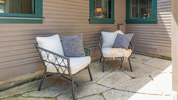 Two wicker chairs with cushions on a stone patio, adjacent to a building with green window frames and horizontal siding.