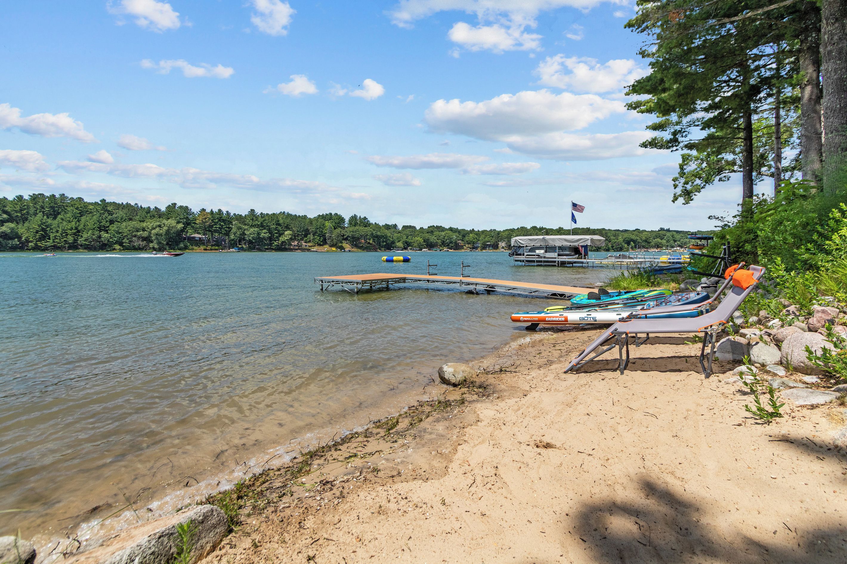 A lakeside beach with clear water, a dock, and a row of lounge chairs.