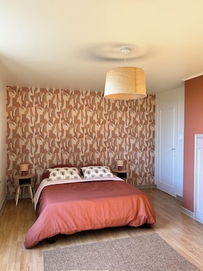 A bedroom with a patterned wallpaper, a bed with a red cover, a hanging lamp, and a door.