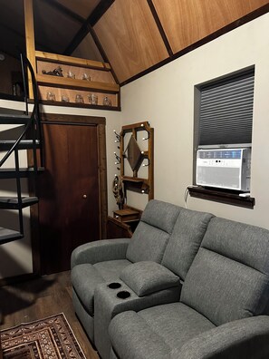 A room with a grey sofa, a wooden staircase, a mirror with lights, and a window with blinds.