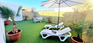 Eine Terrasse mit Kunstgrasanlage, weißen Lounge-Stühlen, einem Tisch mit Stühlen und Topfpflanzen.
