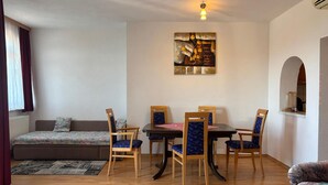Ein Zimmer mit einem Bett, einem Esstisch mit Stühlen, einem Bild an der Wand und einem Fenster mit Vorhängen.