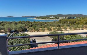 Vista costera desde un balcón con barandilla metálica, con una playa, una pequeña isla y un paisaje montañoso al fondo.