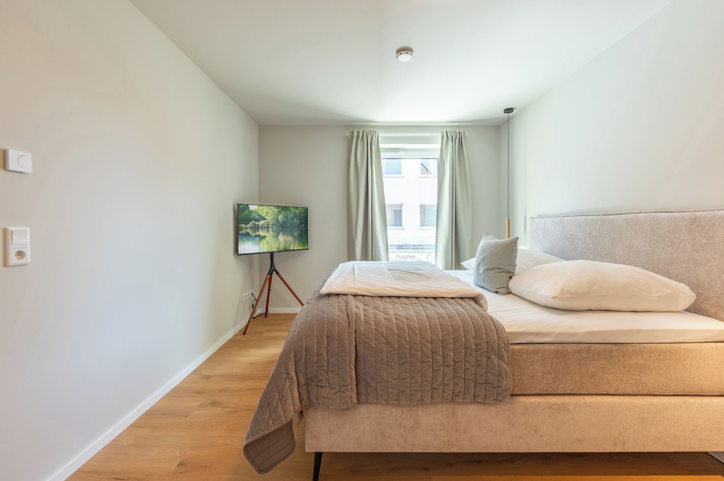 Ein Schlafzimmer mit einem Bett, einem Flachbildfernseher, einem Fenster mit Vorhängen und einer Deckenleuchte.