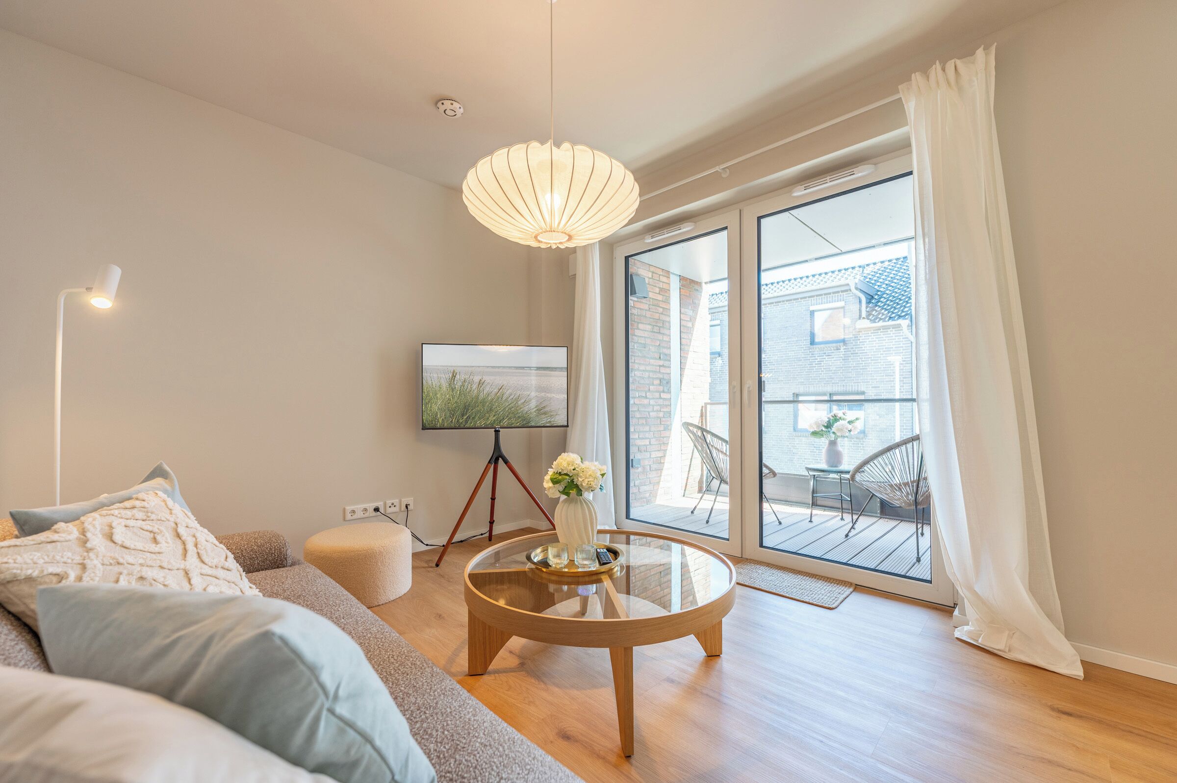Una sala de estar moderna con un sofá, una mesa de centro, un televisor sobre un trípode y vistas a un balcón con mobiliario exterior.