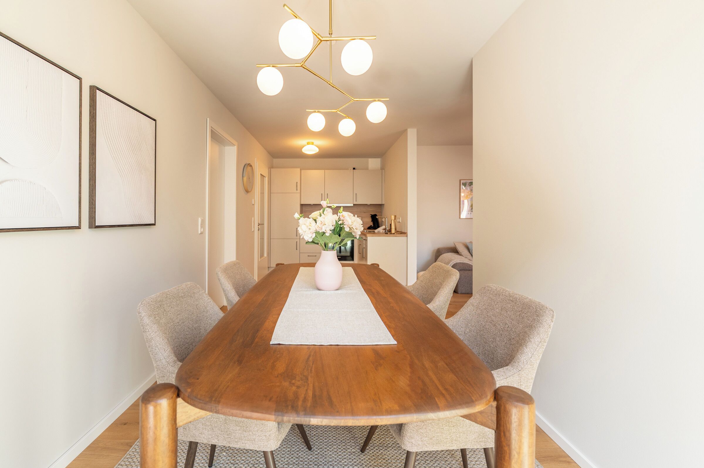 Een moderne eetkamer met een houten tafel, zes gestoffeerde stoelen, een bloevas en een keuken op de achtergrond.