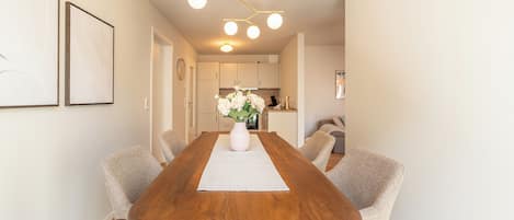 A modern dining room with a wooden table, six upholstered chairs, a vase of flowers, and a kitchen in the background.