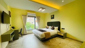 Quarto de hotel com cama, mesa de cabeceira, escrivaninha, cadeira e uma janela grande.