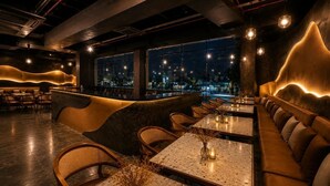 Interior de restaurante com design moderno, apresentando um espaço de bar com assentos e mesas, e vista para a cidade através de grandes janelas.