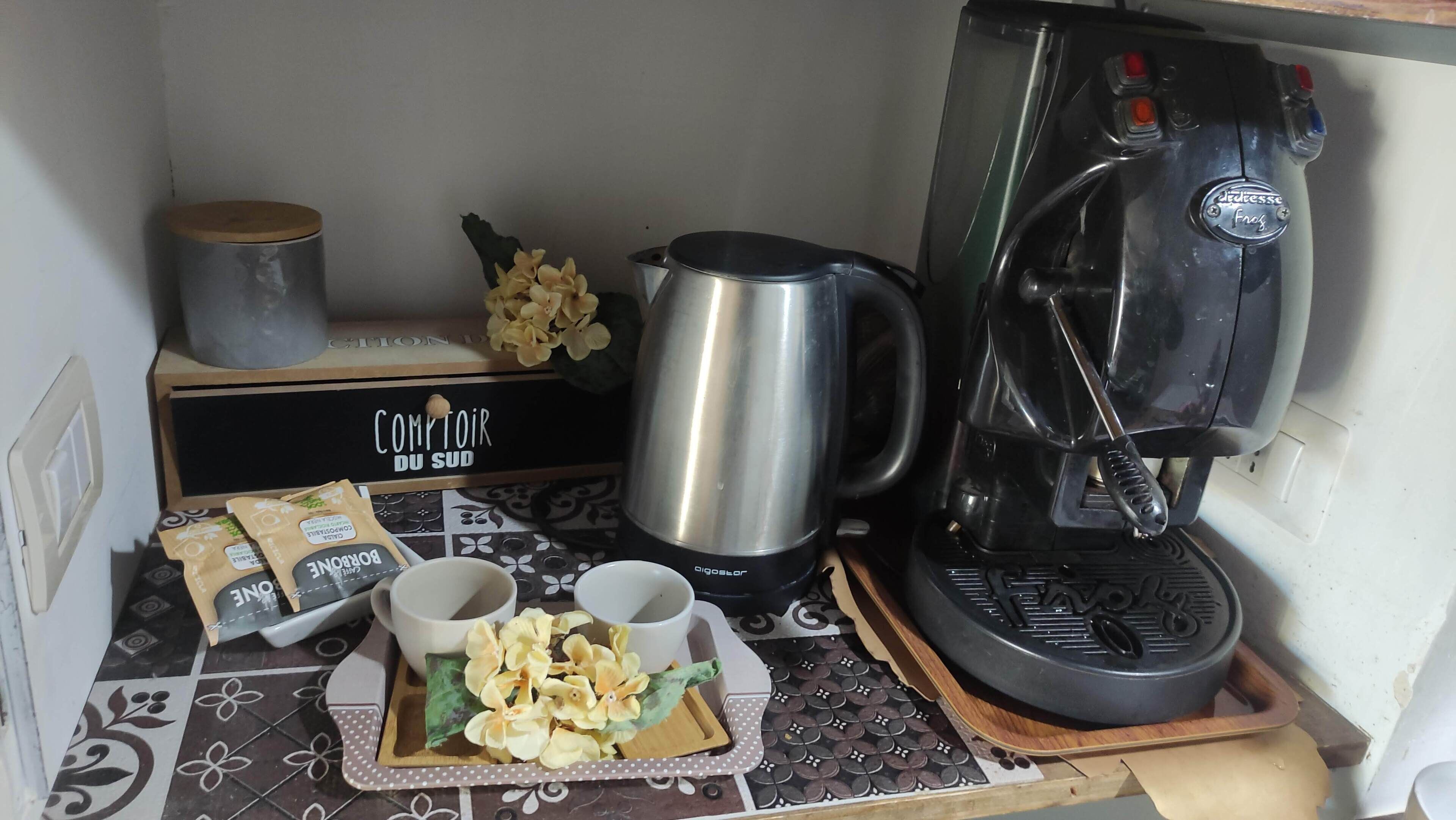 A kitchenette with a coffee maker, a kettle, a box labeled 'Comptoir Du Sud,' and a tray with a bag of biscuits and two cups.