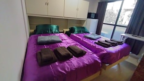 Una habitación con dos camas, cada una con ropa de cama púrpura y toallas marrones. Hay un armario blanco de dos puertas y un televisor de pantalla plana sobre un soporte.