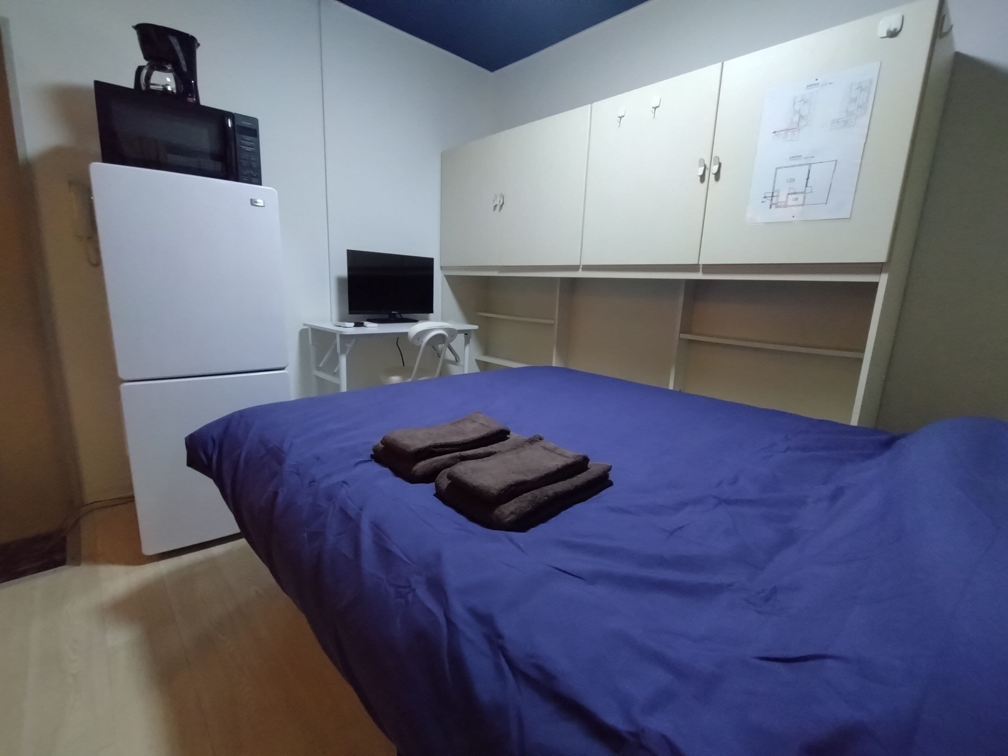 A bedroom with a bed, a desk with a computer, a refrigerator, and a wardrobe.