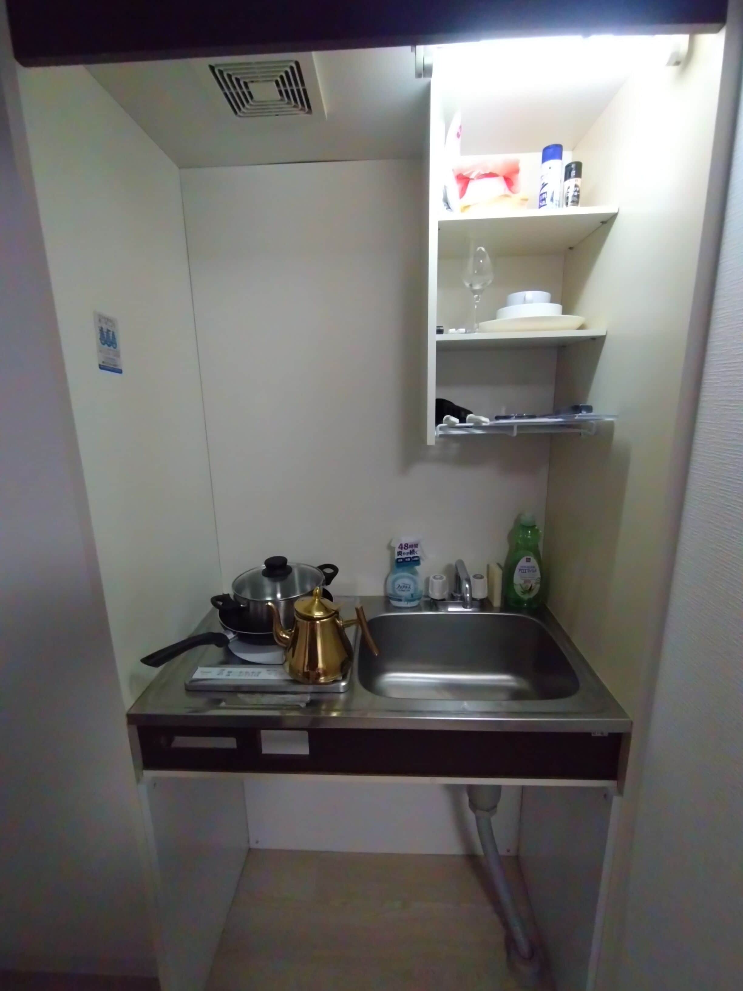 A compact kitchen with a sink, a stove, a kettle, and a small shelf with various items.