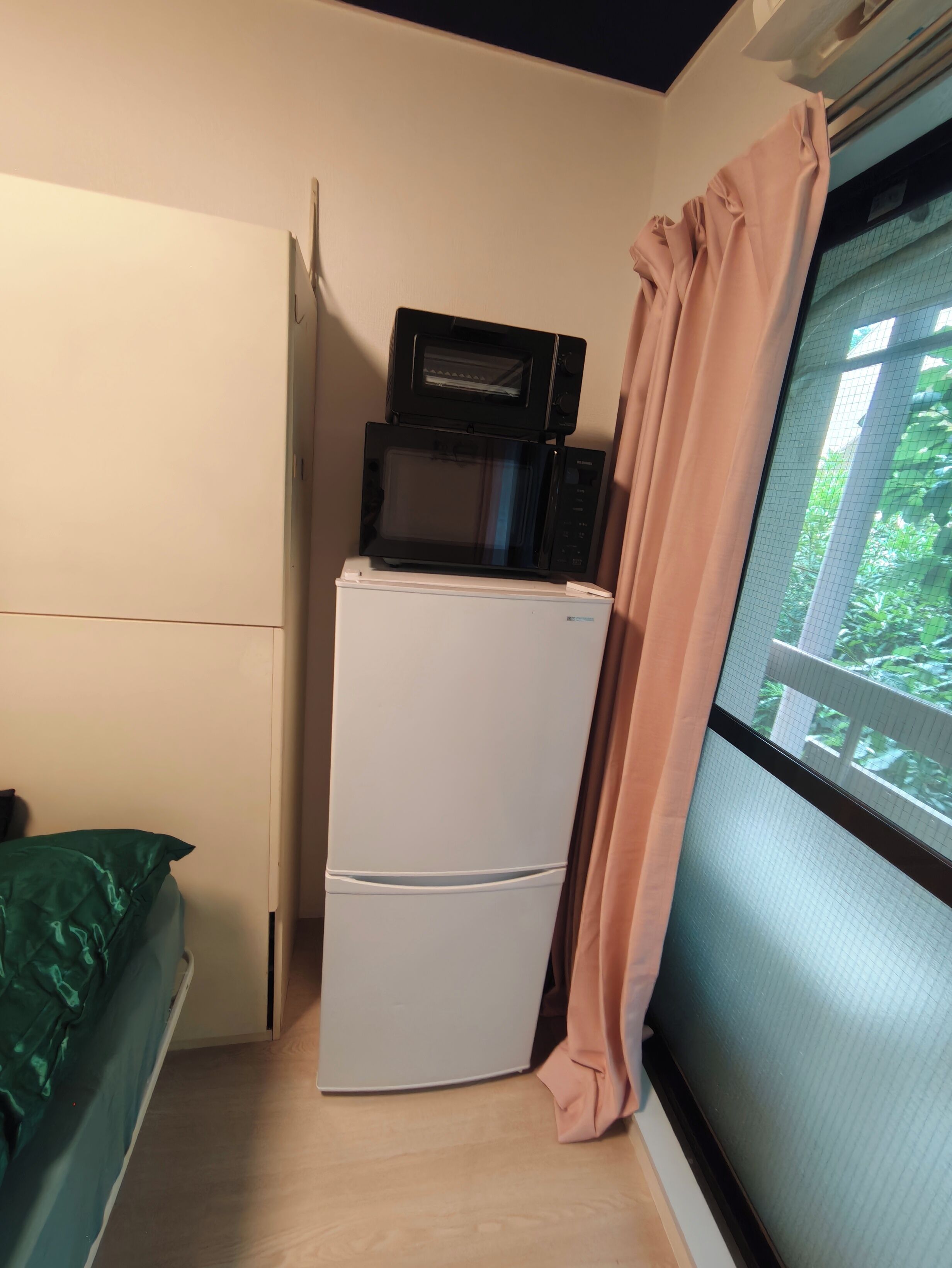 A compact room with a white refrigerator, a microwave on top, a bed with a green cover, and a window with a view of greenery.