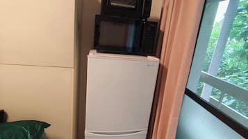 A compact room with a white refrigerator, a microwave on top, a bed with a green cover, and a window with a view of greenery.
