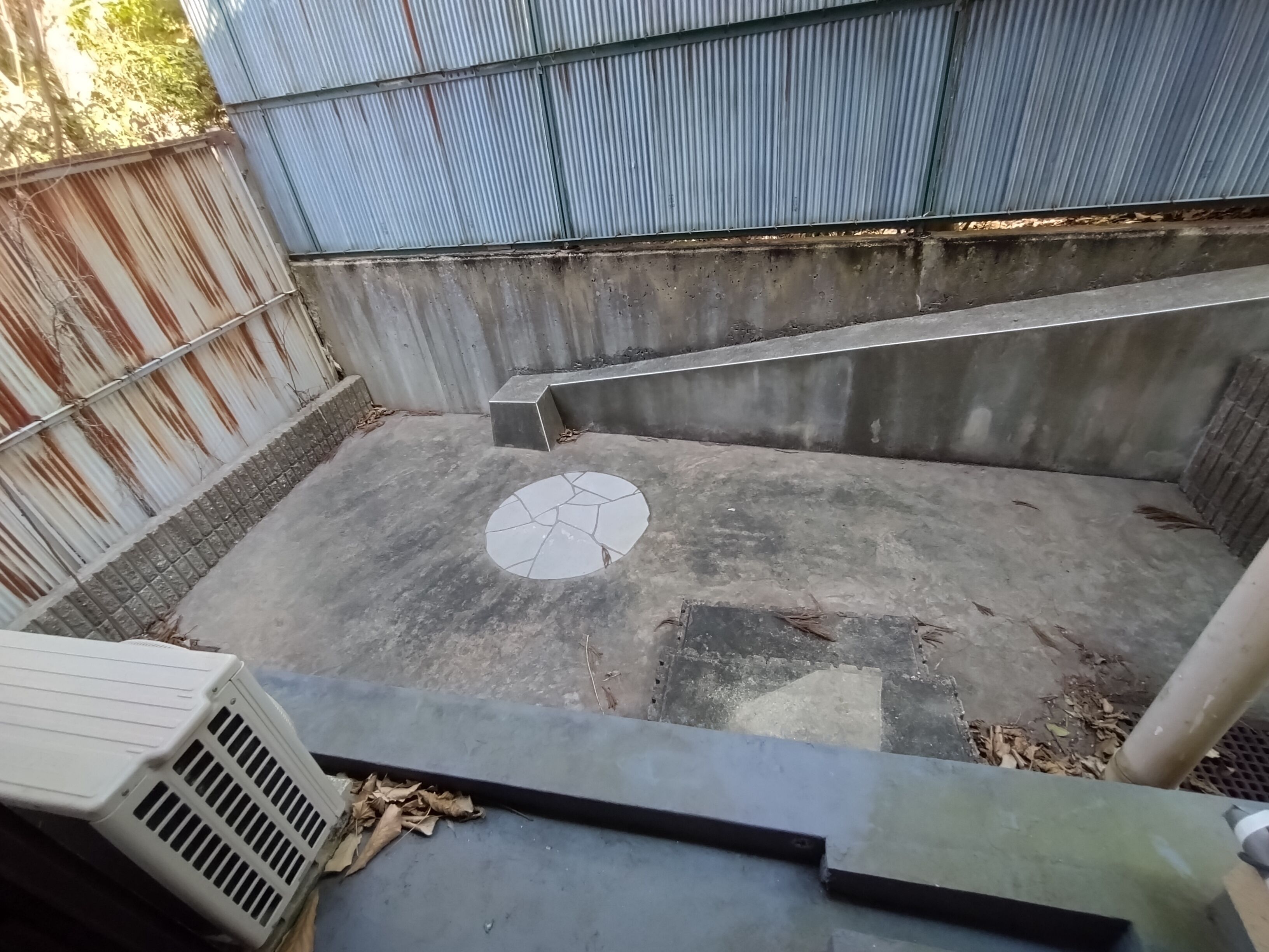 A concrete outdoor area with a metal fence, a tiled floor with a circular pattern, and a concrete bench.