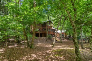 A cabin with a wooden deck and multiple levels, surrounded by a forested area. - Riverfront Cabin: Hot Tub, Sauna, Arcade & Views (Ellijay)