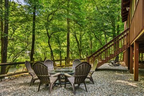 An outdoor seating area with a table and chairs, surrounded by a wooden deck and a forested backdrop. - Riverfront Cabin: Hot Tub, Sauna, Arcade & Views (Ellijay)