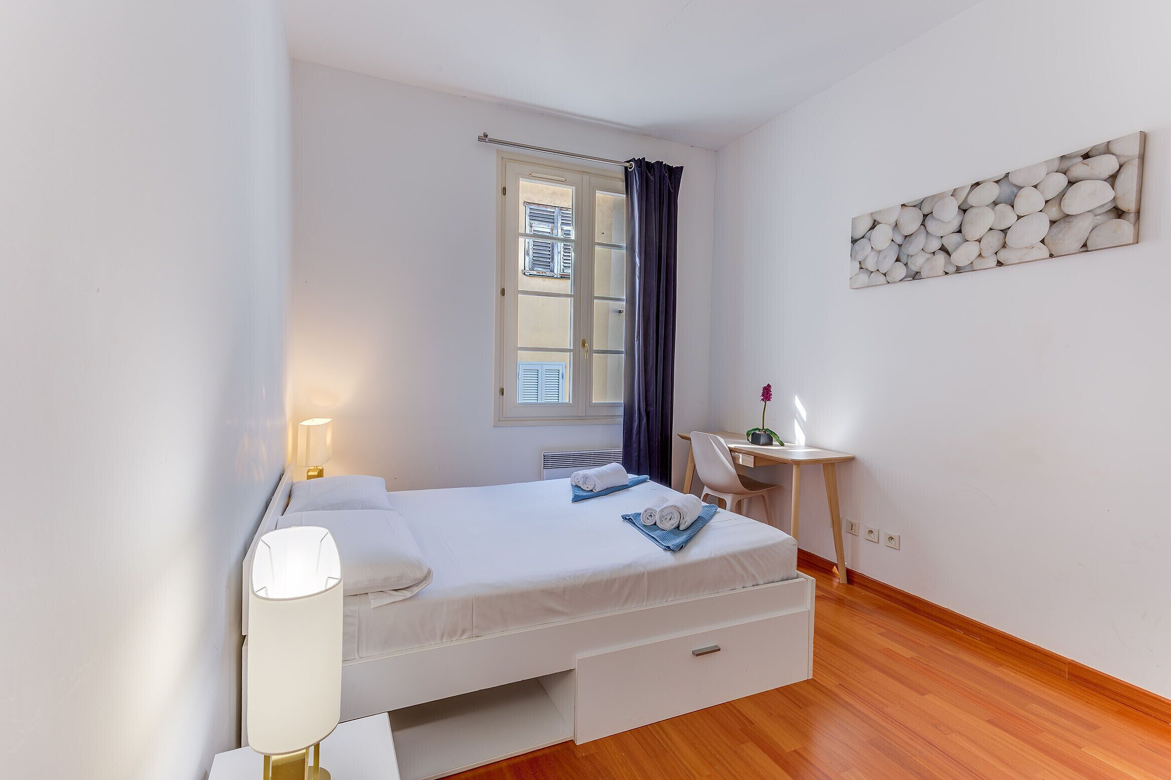 A bedroom with a bed, bedside table, lamp, window with curtains, and a desk with a chair.