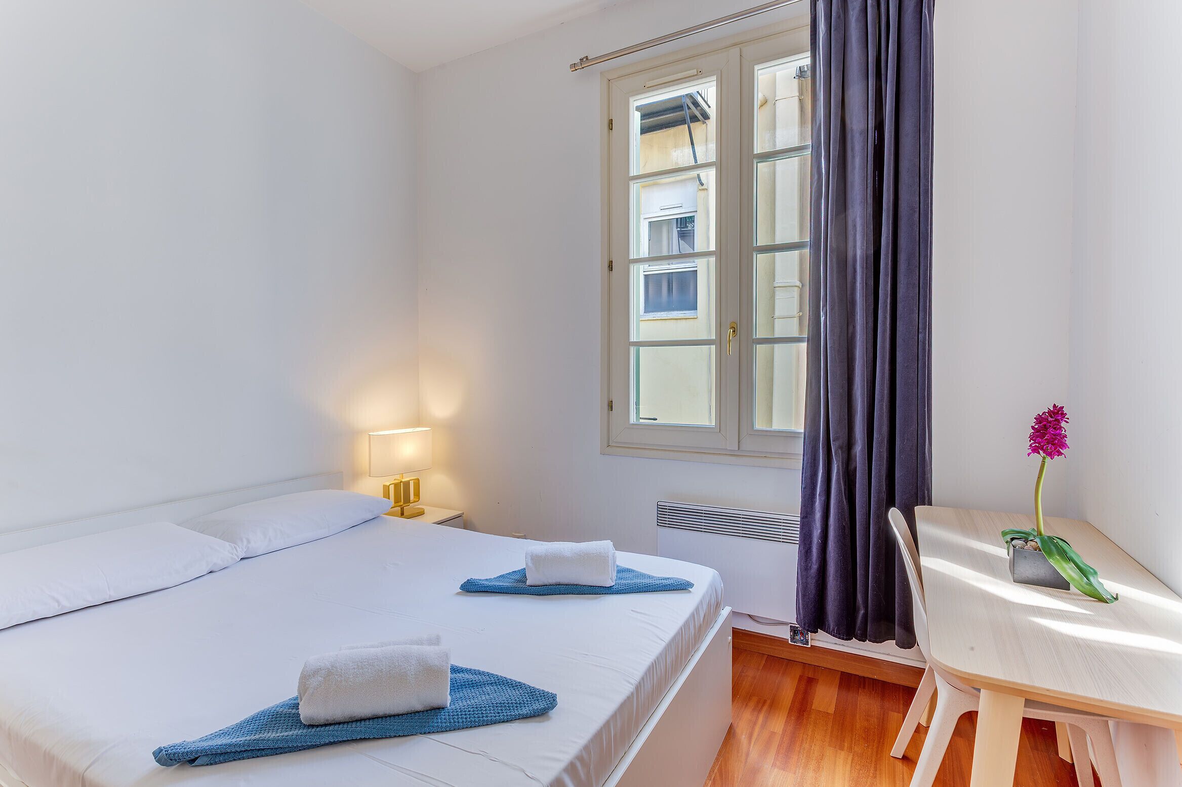 Ein Hotelzimmer mit zwei Betten, einem Fenster mit Vorhängen, einem Nachttisch mit Lampe und einem kleinen Tisch mit Pflanze.