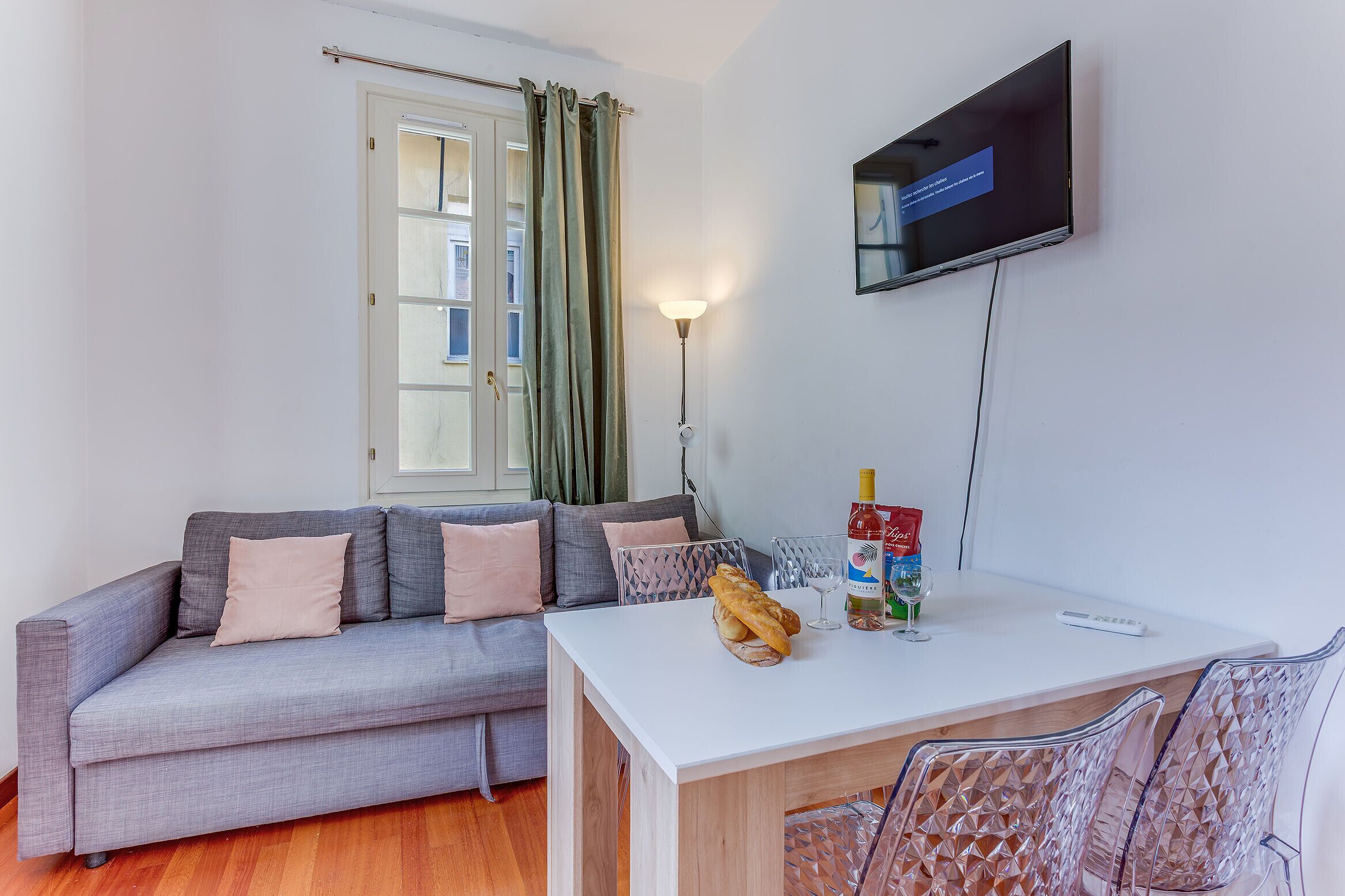 A living room with a sofa, a dining table, a TV mounted on the wall, and a window with curtains.