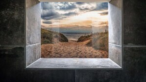 Une vue encadrée d’une plage de sable avec des dunes, que l’on aperçoit à travers une fenêtre dont le cadre est en béton.