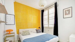 Un dormitorio con cama, mesita de noche, lámpara, ventana con cortinas y un cuadro en la pared.