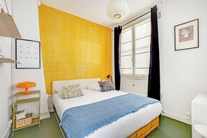 Ein Schlafzimmer mit einem Bett, einem Nachttisch, einer Lampe, einem Fenster mit Vorhängen und einem Wandbild.