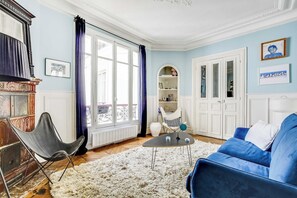 Ein Wohnzimmer mit einem blauen Sofa, einem schwarzen Sessel, einem weißen Ohrensessel, einem kleinen runden Tisch und einem weißen, flauschigen Teppich.