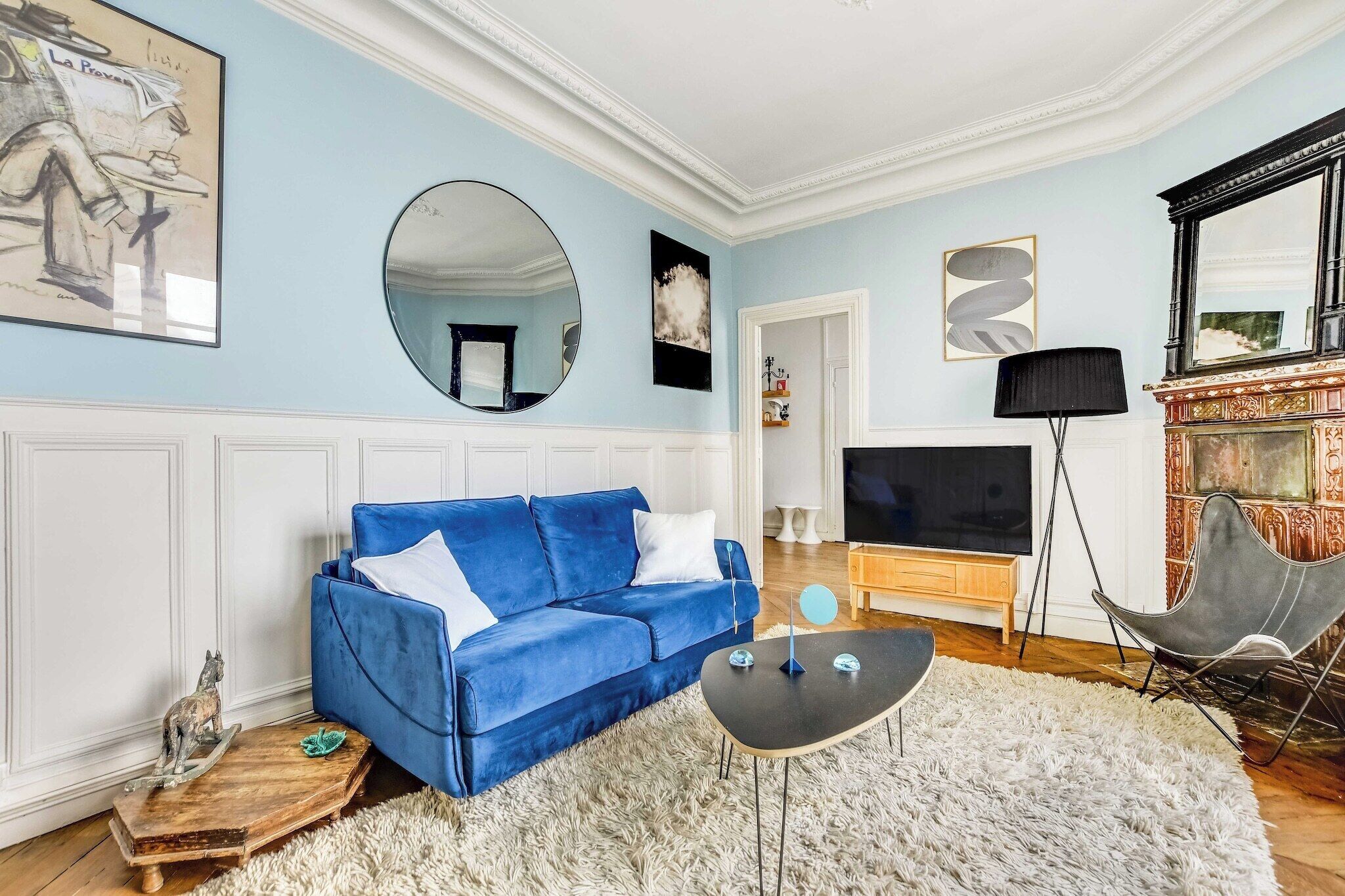 Un salon avec un canapé bleu, un miroir rond, un téléviseur à écran plat et une cheminée décorative.