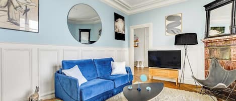 Una sala de estar con un sofá azul, un espejo redondo, un televisor de pantalla plana y una chimenea decorativa.