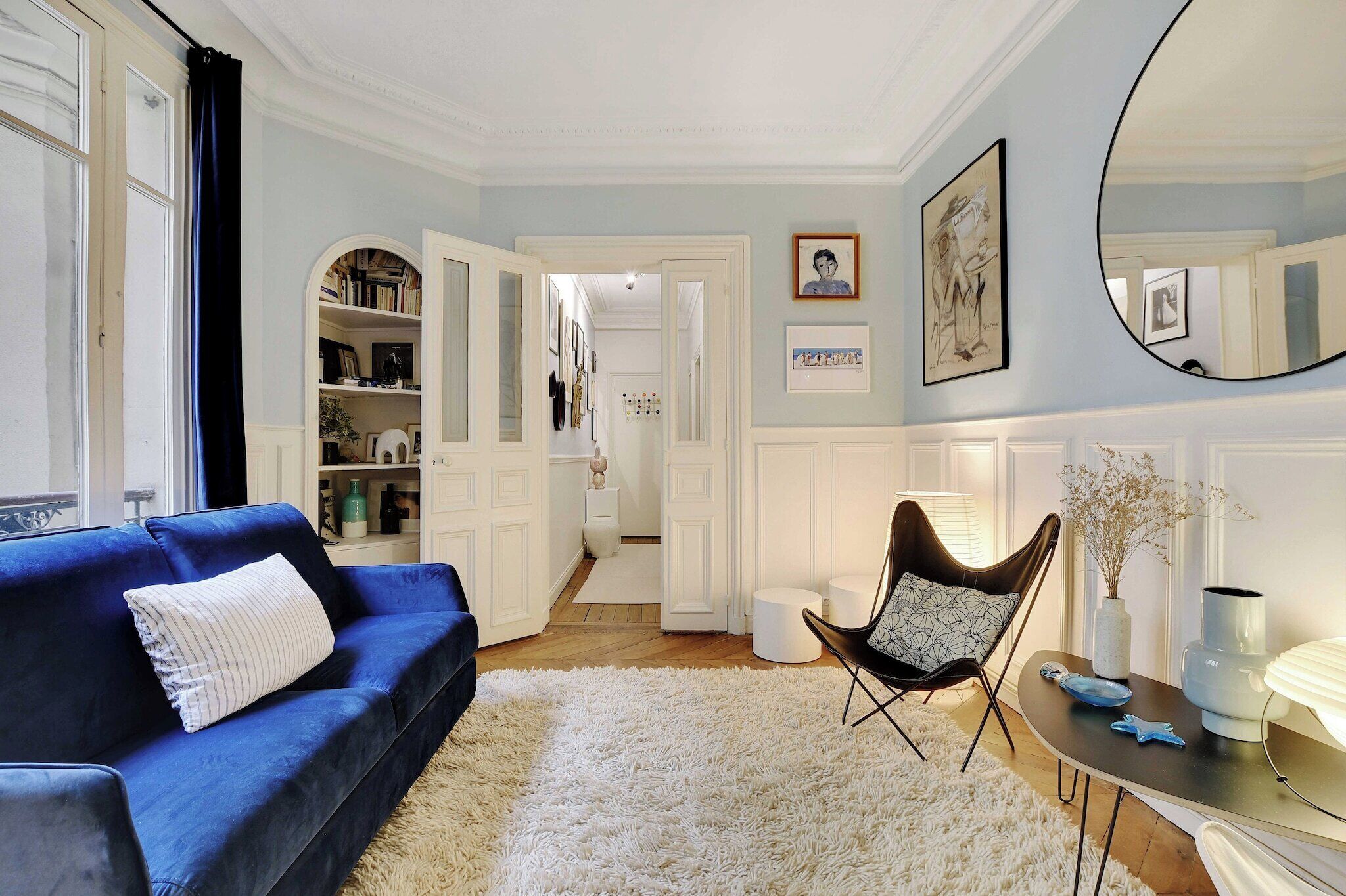 A living room with a blue sofa, a white shag rug, a black chair, and a round mirror on the wall.