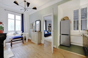 Ein Zimmer mit Esstisch, Stühlen, einer Bank, einem Spiegel, einem Kleiderschrank, einem Bett, einem Kühlschrank und einem Fenster.