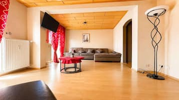 Ein Wohnzimmer mit einer Couch, einem roten Tisch, einer Stehlampe und einem an der Wand befestigten Fernseher.