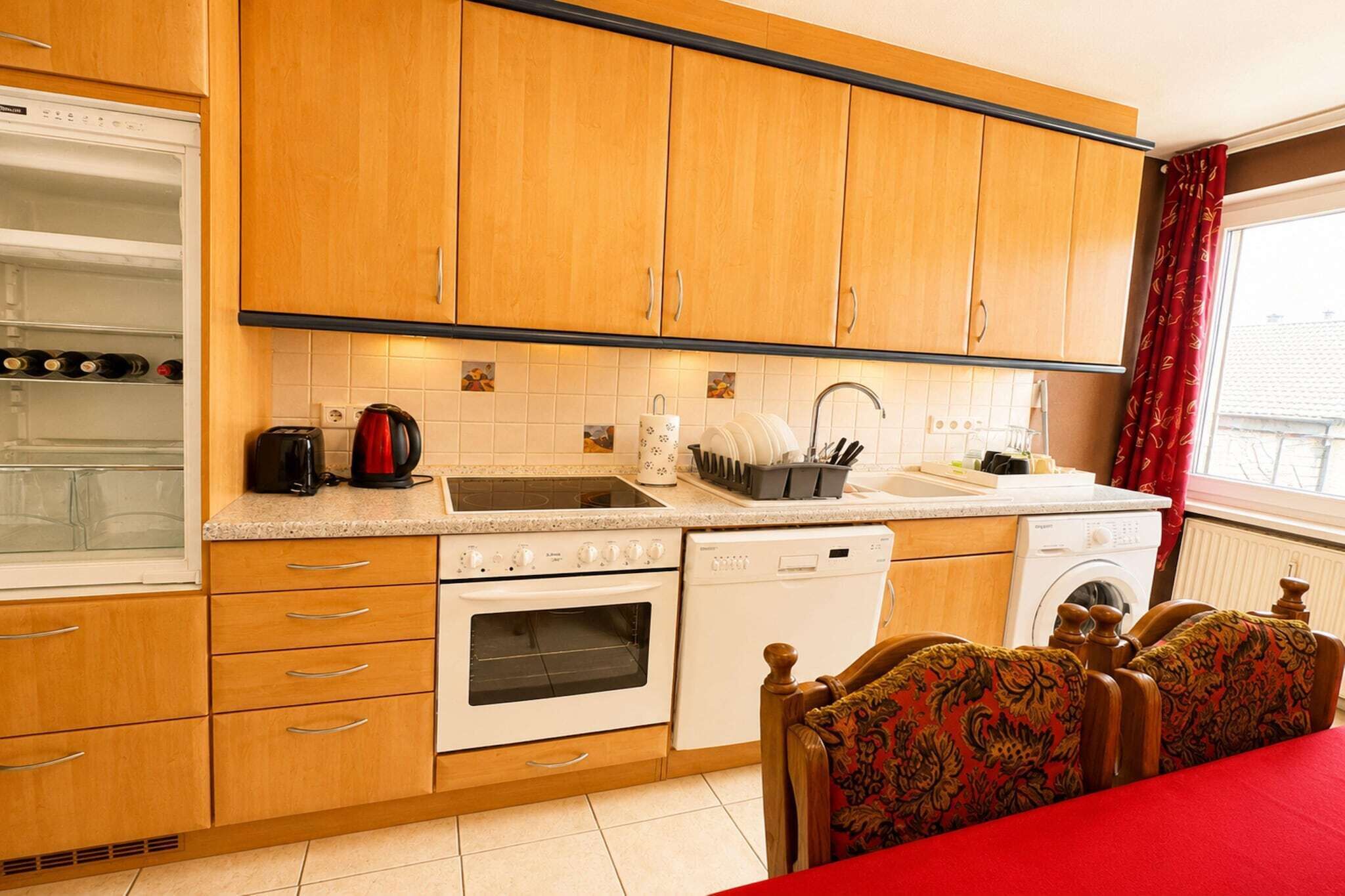 A compact kitchen with wooden cabinets, a built-in oven, a dishwasher, and a dining area with a red table and patterned chairs.