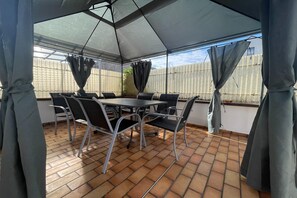 Ein überdachter Patio-Bereich mit Tisch und Stühlen, einem gefliesten Boden und Vorhängen.
