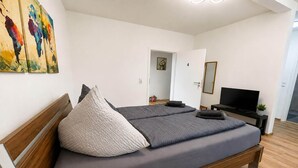 Un dormitori modern amb un llit de fusta, roba de llit gris i un televisor sobre un suport.