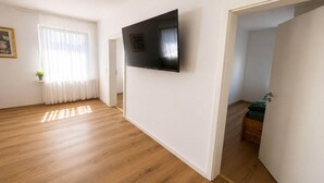 Une pièce dotée d’un téléviseur à écran plat fixé au mur, d’un plancher en bois, d’une fenêtre ornée de rideaux légers et d’une porte donnant sur une autre pièce.