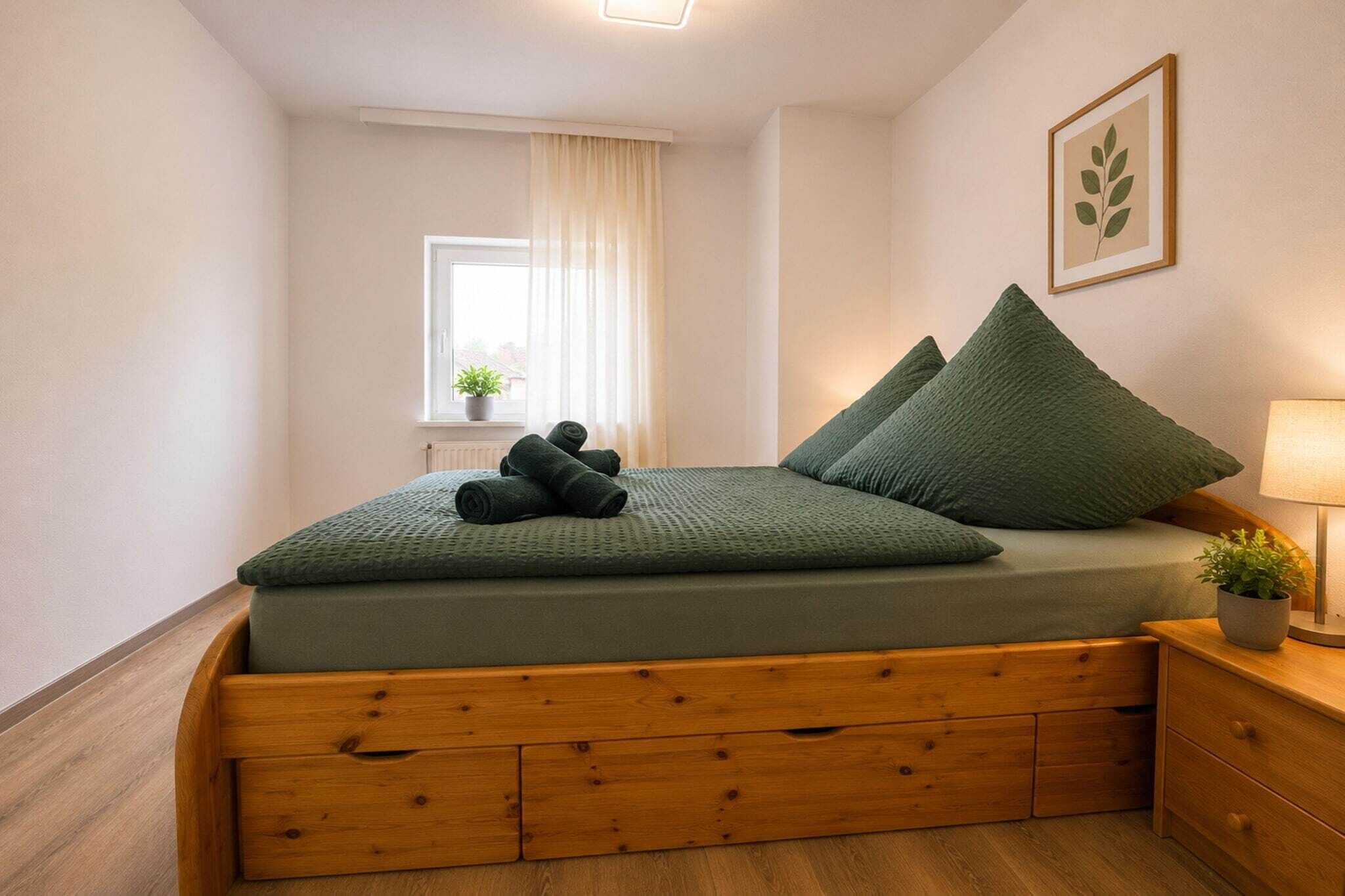 A neatly made bed with a green bedspread and pillows, a wooden nightstand with a lamp, a window with curtains, and a framed plant on the wall.