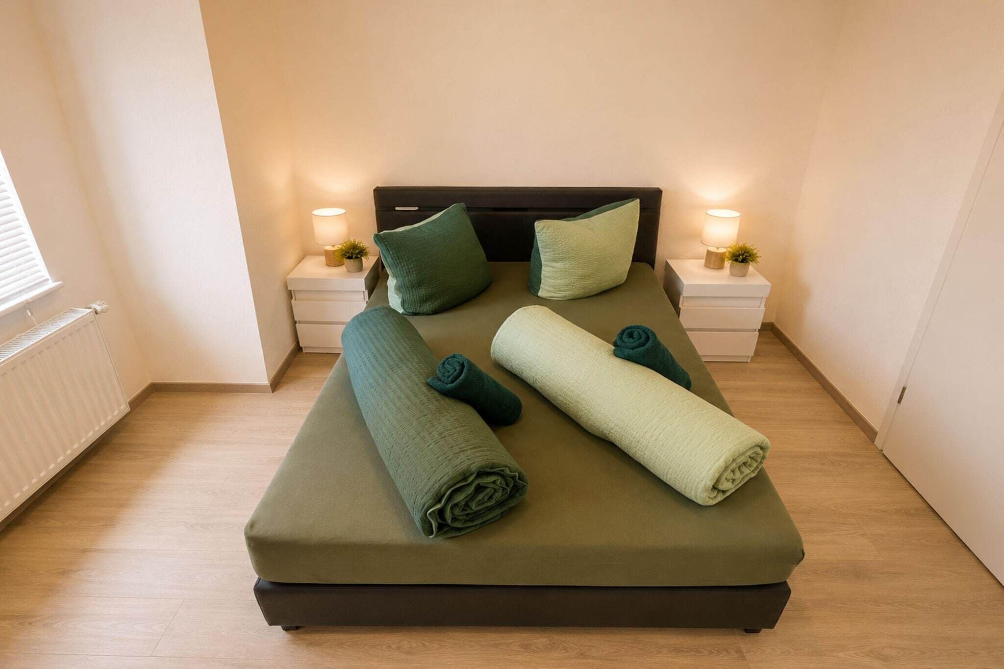 A neatly made bed with pillows and rolled towels, two bedside tables with lamps, and a radiator on the wall.