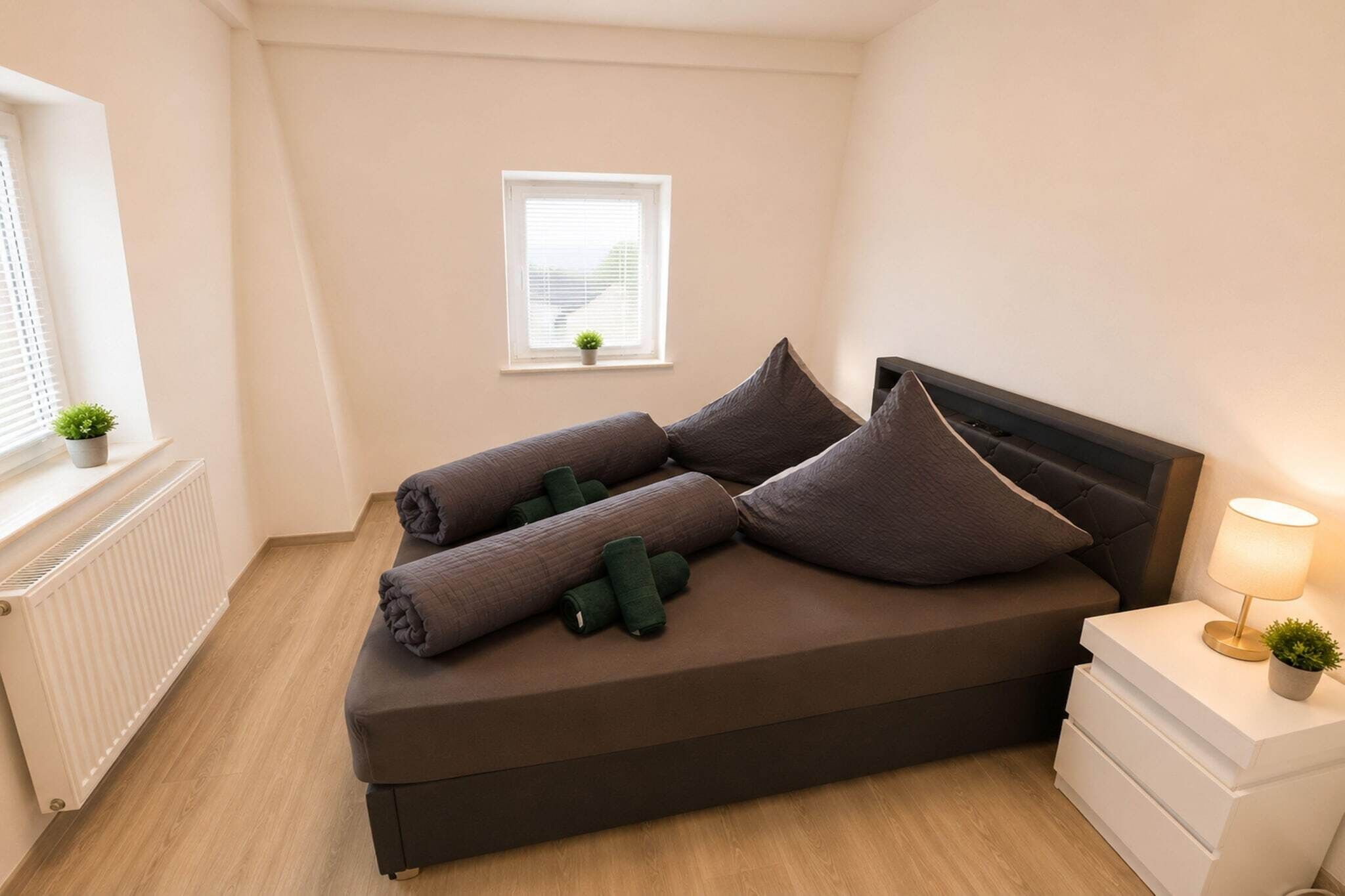 A bedroom with a bed, a bedside table with a lamp, a window with blinds, and a small plant on the windowsill.
