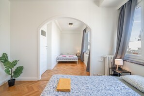 A hotel room with a bed, a bedside table with a lamp, a potted plant, and a view through a window. - Comfortable 3BHK Aprt for up to 6 Guests (Antwerp)