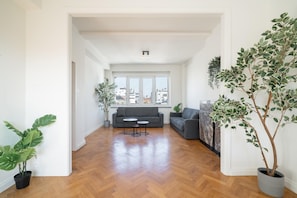 A spacious living area with a wooden floor, a grey sofa, and two potted plants. - Bright & Spacious 3BHK Apartment (Antwerp)