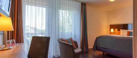 A hotel room with a bed, a chair, a desk with a lamp, and a window with curtains.