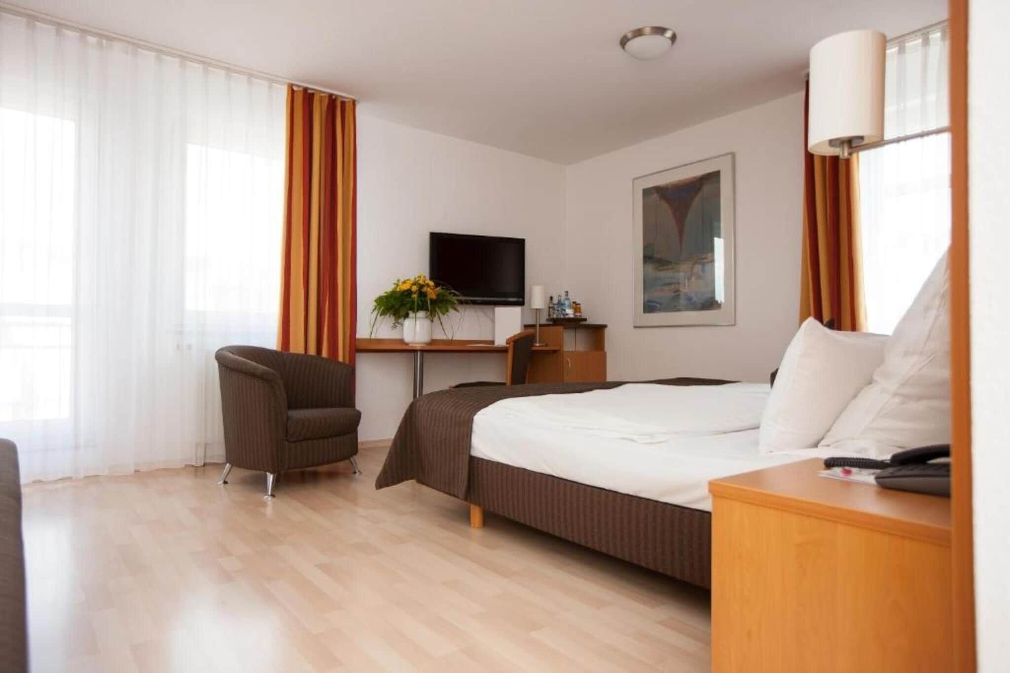 Una habitación de hotel con cama, escritorio, silla, televisor y ventana con cortinas.