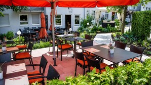 En udendørs patio med borde og stole, hvoraf nogle har orange hynder, og en stor rød parasol giver skygge.